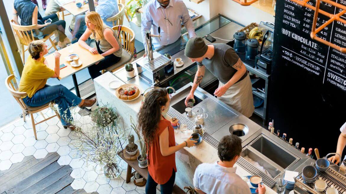 Restaurant Marketing Tactics Busy café with baristas preparing drinks behind the counter and customers engaging at tables, illustrating effective restaurant marketing tactics that enhance in-store experience and customer retention.