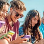 Group of young adults sitting outdoors on a sunny day, smiling and looking at their smartphones, wearing colourful clothes and sunglasses, symbolising digital engagement and social connection.